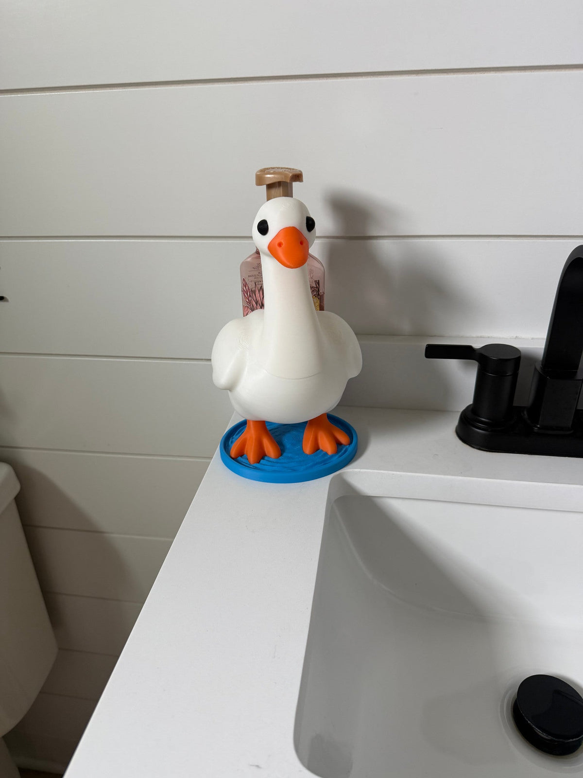 Goose Soap Dispenser Holder — a white goose-shaped holder with an orange beak and feet, standing on a blue base beside a bathroom sink.