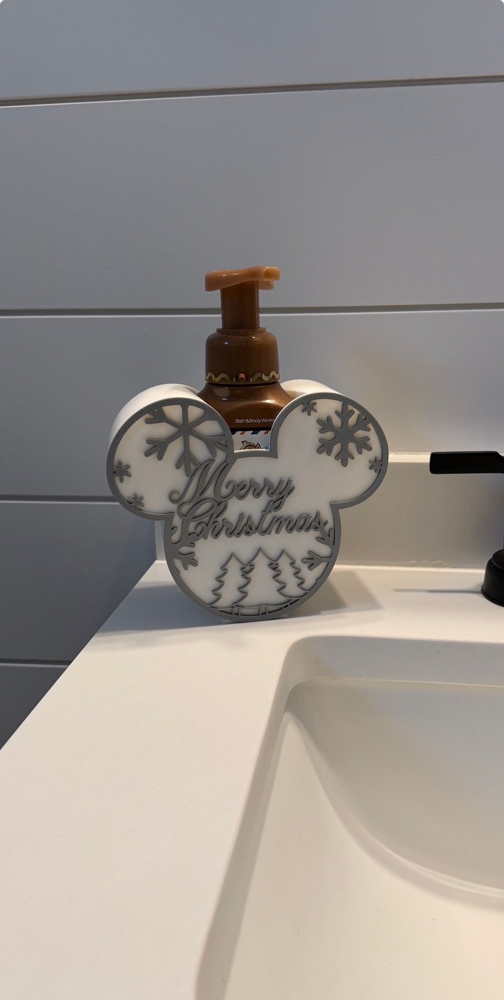 Merry Christmas Soap Dispenser Holder ā a white rounded holder with ear shapes, snowflake and tree detailing and 'Merry Christmas' lettering, holding a pump soap bottle at a sink.