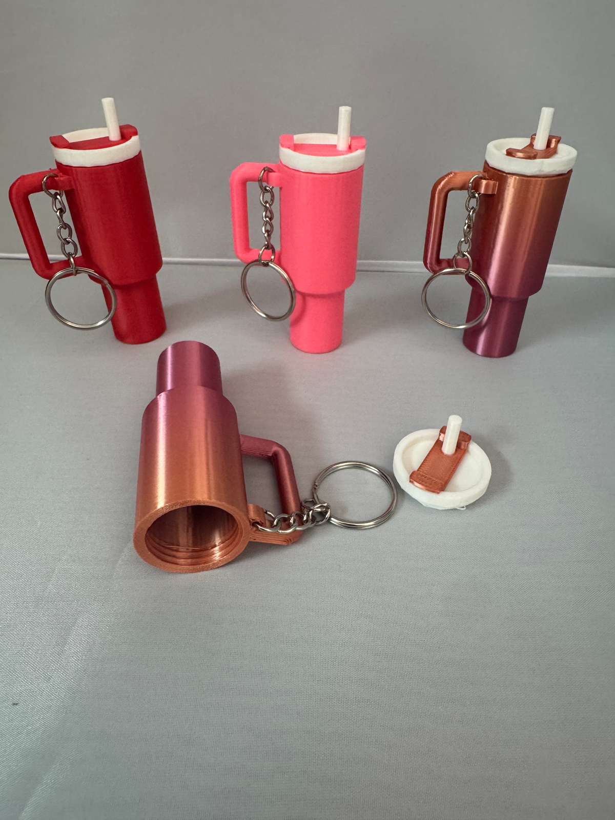 Mini Tumbler Lip Balm Holder ā four miniature tumbler-shaped keychains in red, pink, copper and rose, each with a handle, lid and straw, with one shown open to reveal the lip balm compartment.
