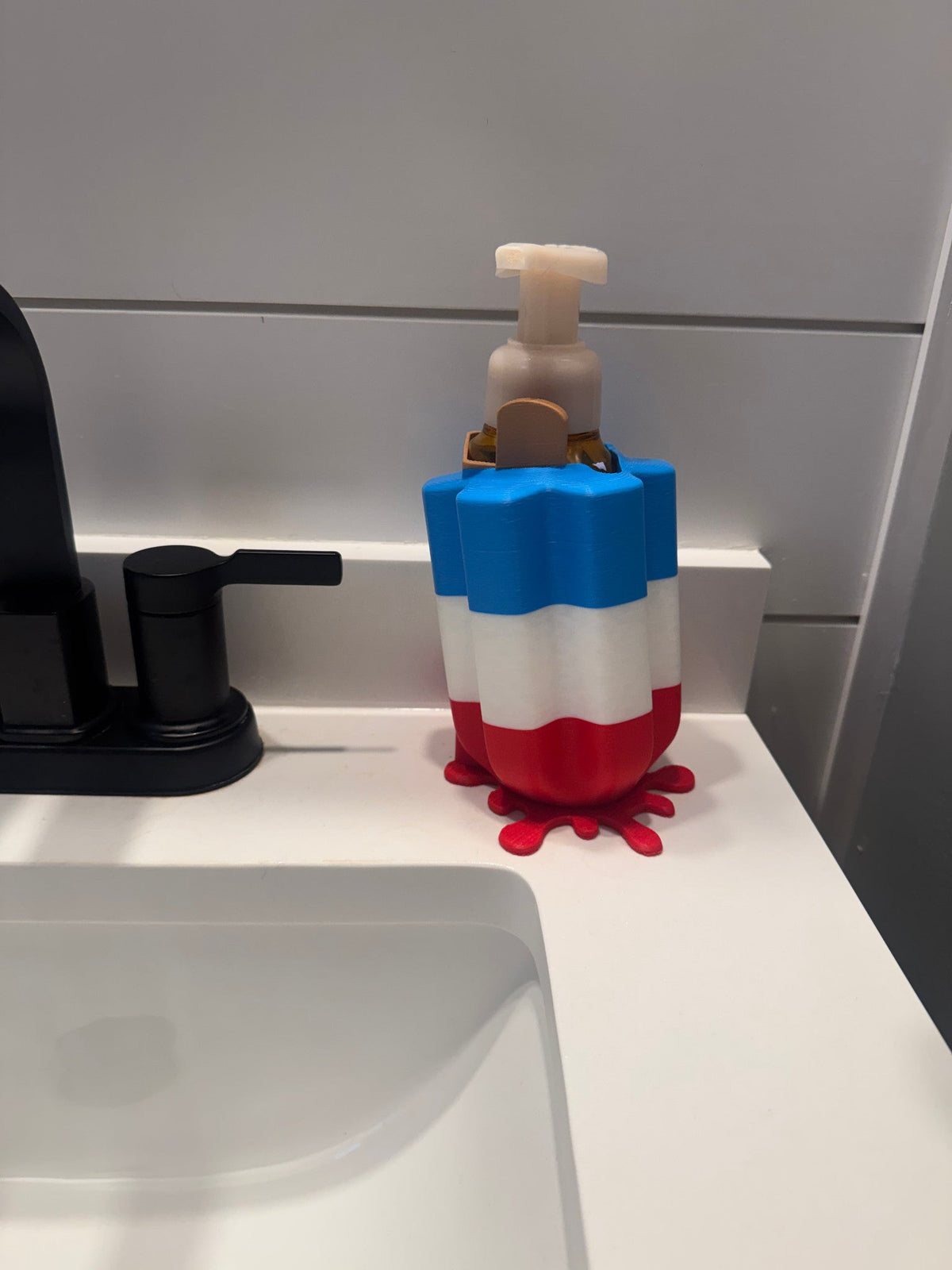 Patriotic Popsicle Soap Dispenser Holder β a red, white and blue striped popsicle-shaped holder with a red drip base, cradling a foaming soap bottle at a bathroom sink.