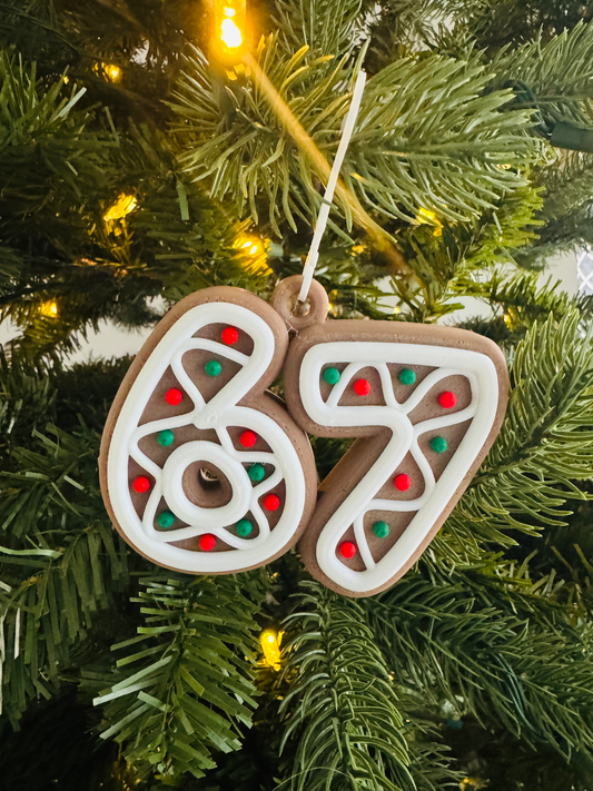 "67" Gingerbread Ornament — a gingerbread-style ornament shaped as the number 67, outlined in cream icing with red and green candy dots, hanging on a lit Christmas tree.