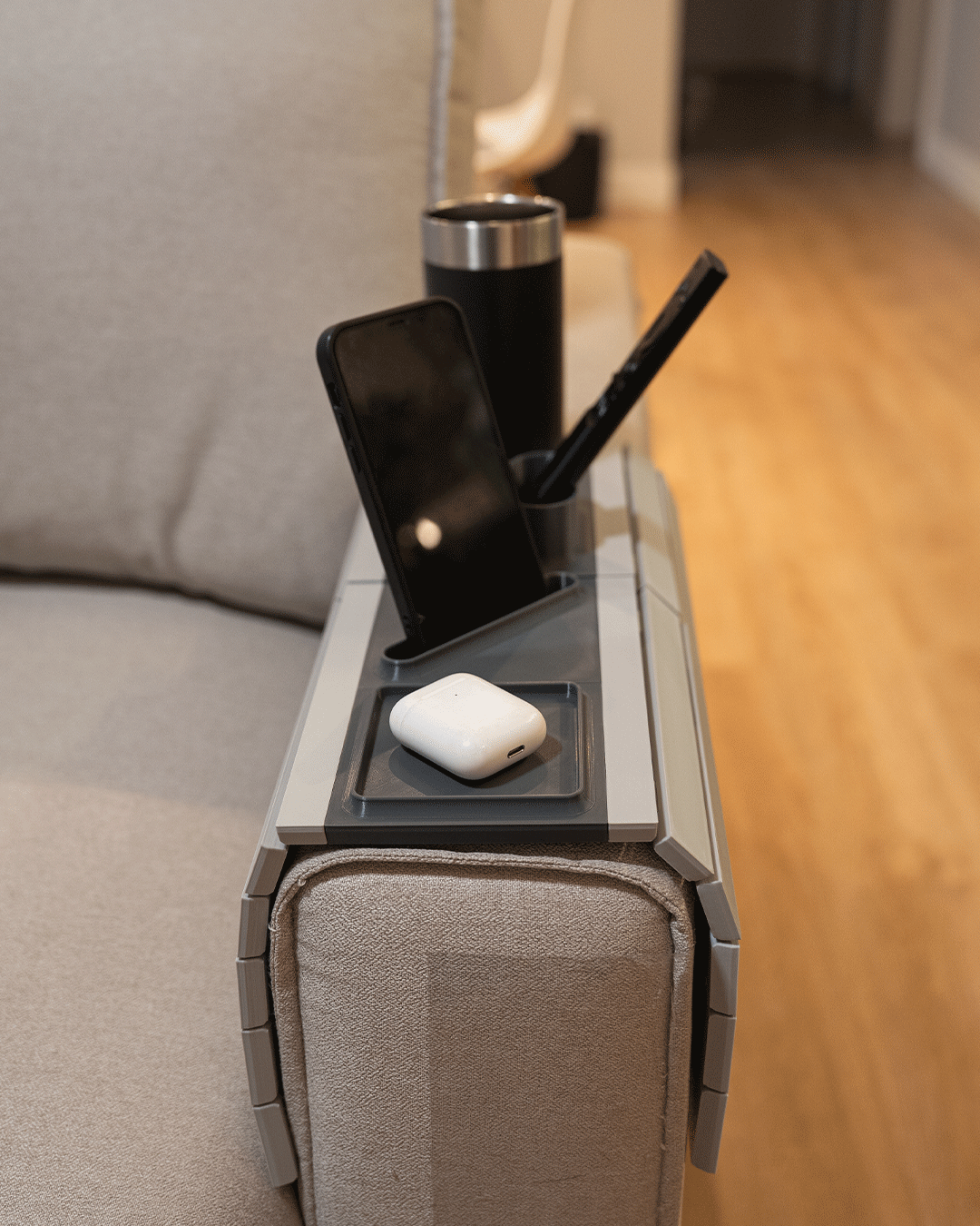 All In — Modular Sofa Side Table — close view on a beige sofa arm holding a black tumbler, an upright phone, a stylus and an earbuds case.