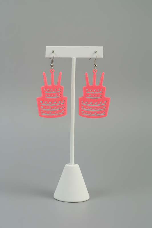 Birthday Cake Earrings (Pink) — bright pink cake-shaped dangle earrings hanging from silver hooks, on a soft light-gray studio background.