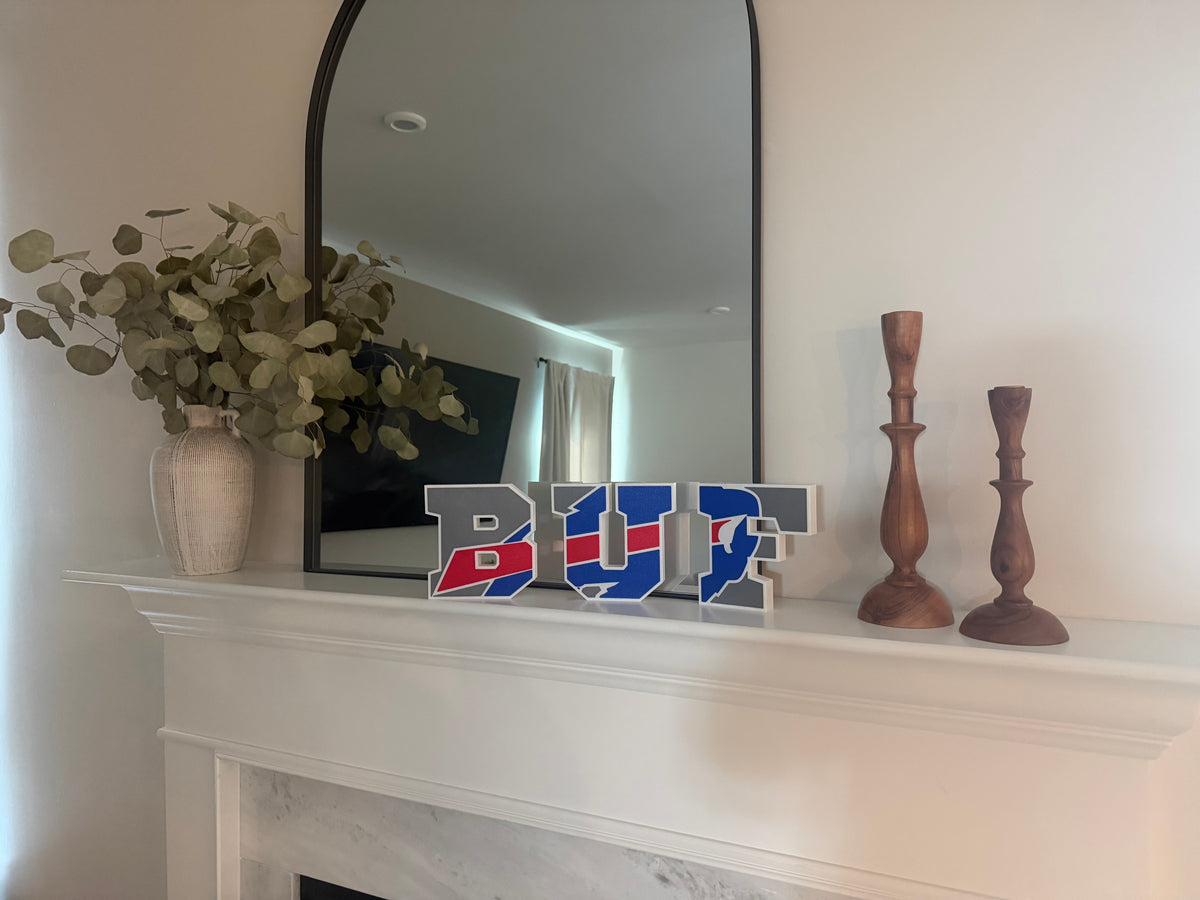 Buffalo Football Varsity Letter Sign — three free-standing layered pieces in blue, red and gray, standing on a white fireplace mantel in front of an arched mirror.