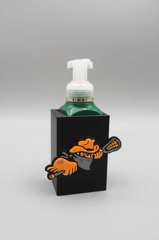 Buffalo Lacrosse Soap Dispenser Holder — a black sleeve-style holder with a raised front emblem, cradling a green foaming soap bottle with a clear pump, on a soft light-gray studio background.