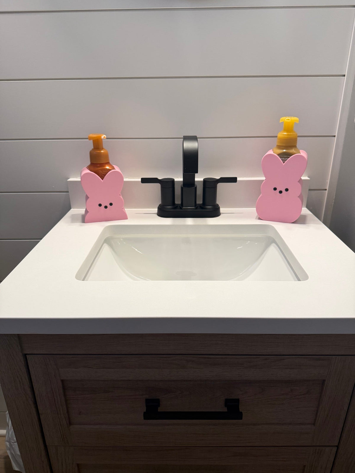 Bunny Soap Dispenser Holder — a pair of pink marshmallow-bunny shaped holders with simple faces, flanking a black faucet on a white vanity.