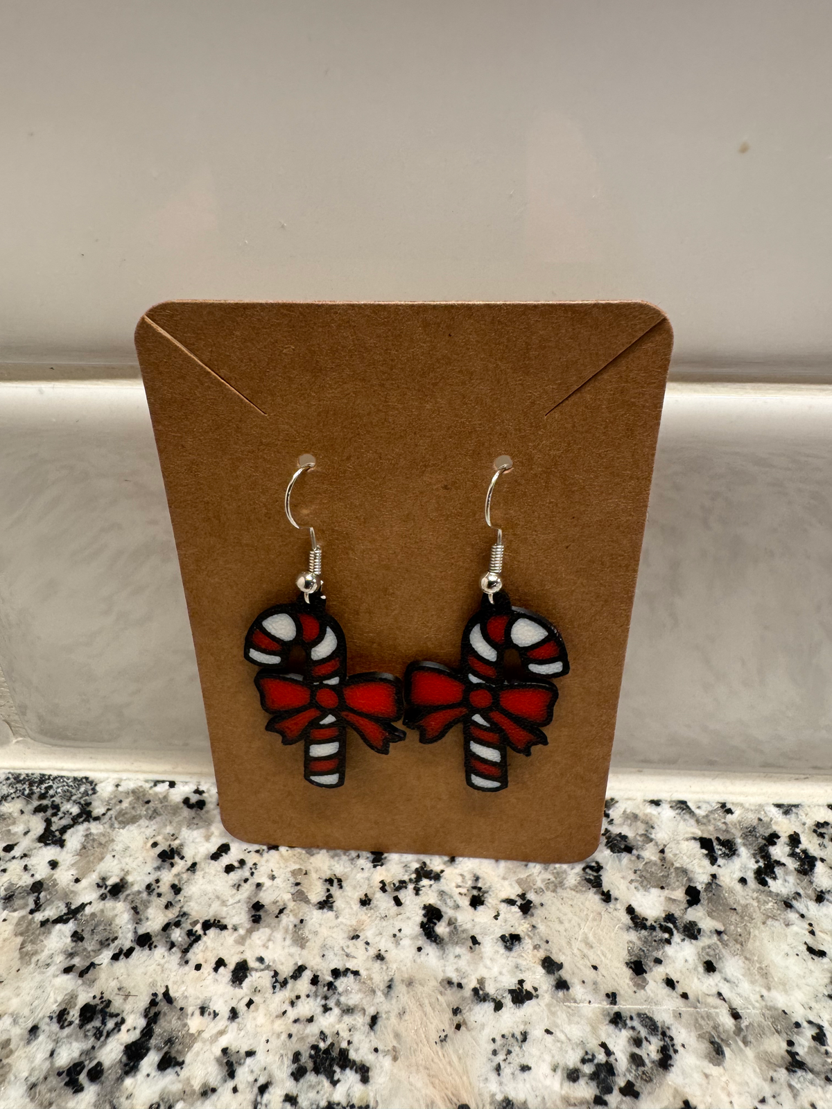 Candy Cane Dangle Earrings — a pair of red and white striped candy cane earrings with red bows, on silver hooks, displayed on a kraft card.