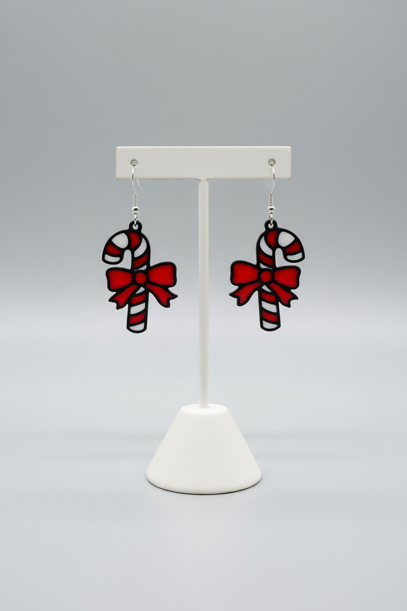 Candy Cane Dangle Earrings — a pair of red and white striped candy cane earrings with black outlines and red bows, hanging from silver hooks, on a soft light-gray studio background.