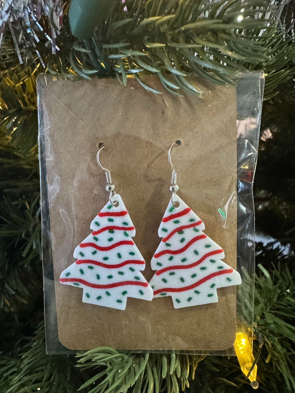 Christmas Tree Snack Cake Earrings — a pair of green tree-shaped dangle earrings with white icing stripes and coloured sprinkles, on silver hooks.