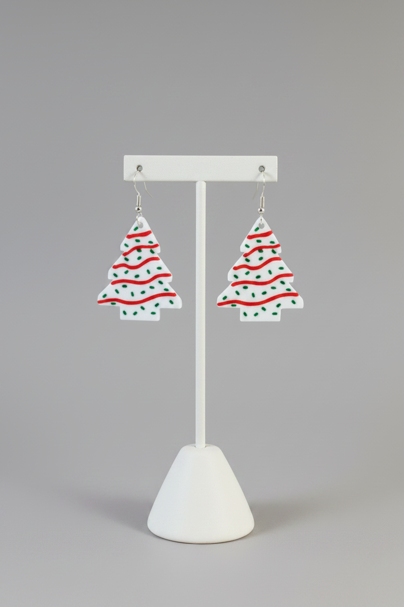 Christmas Tree Snack Cake Earrings — a pair of Christmas tree-shaped dangling earrings with a white base, red wavy stripes, and green dots, on a soft light-gray studio background.