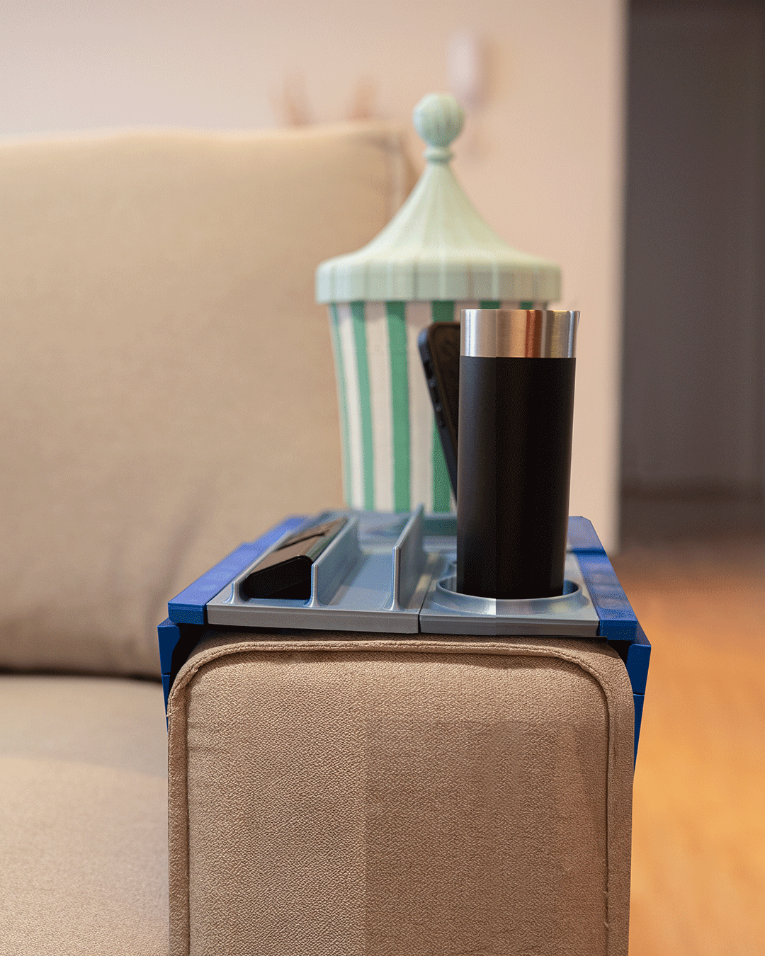Couch Buster — Modular Sofa Side Table — a blue-edged tray on a tan sofa arm holding a tumbler, beside a striped lamp.