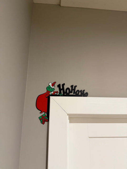 Green Grump Corner Door Topper — a green character figure with a red sack and a gift, mounted in the top corner of a white door frame beside raised lettering.