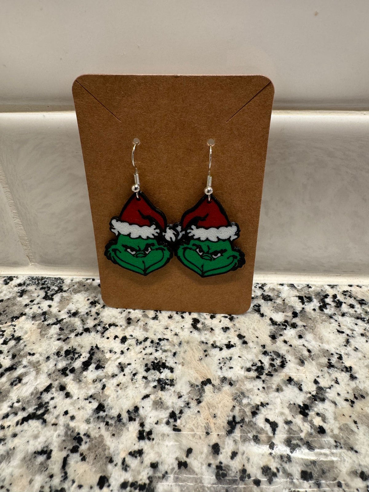 Green Grump Dangle Earrings — a pair of green character-face earrings in red and white santa hats, on silver hooks, displayed on a kraft card.