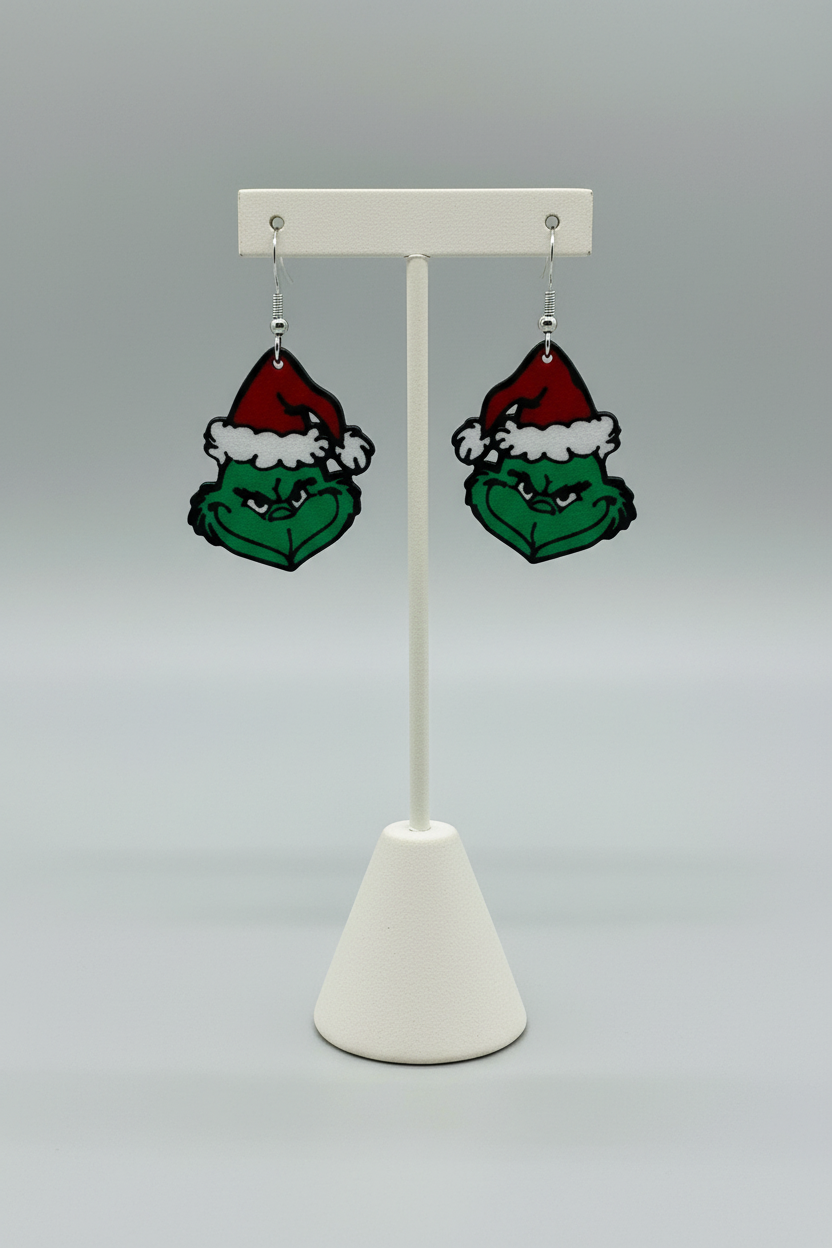 Green Grump Dangle Earrings — a pair of green character-face dangle earrings in red and white hats, on silver hooks, on a soft light-gray studio background.
