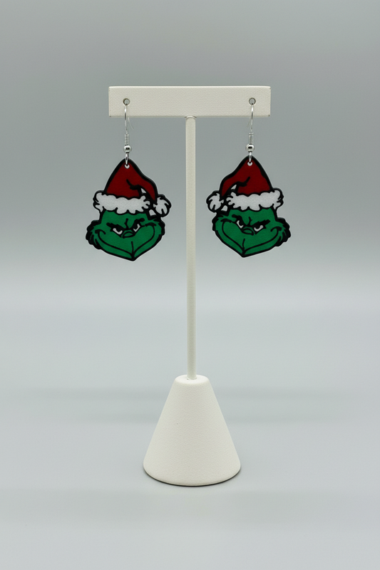 Green Grump Dangle Earrings — a pair of green character-face dangle earrings in red and white hats, on silver hooks, on a soft light-gray studio background.