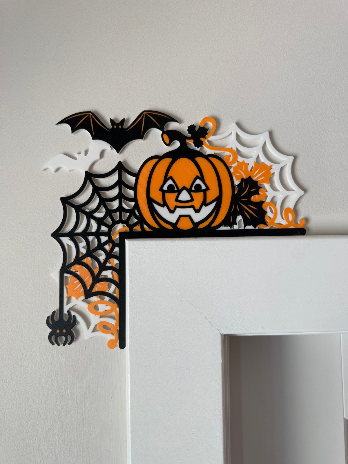 Jack-o'-Lantern & Spiderweb Corner Door Topper — a black and orange corner piece with a spiderweb, a carved pumpkin, a bat and a dangling spider, on a white door frame.