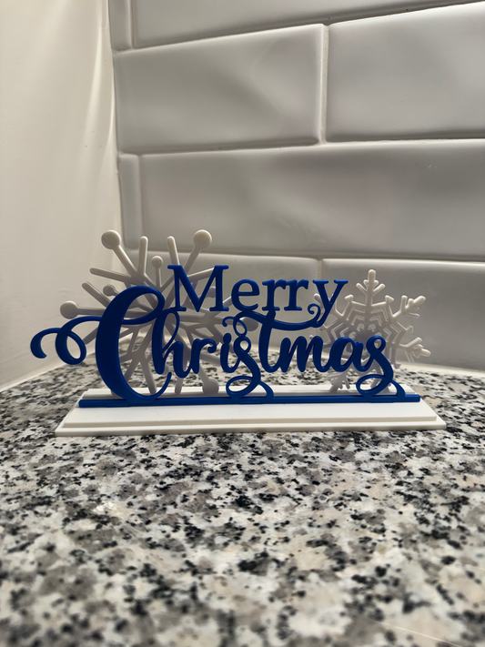Layered Merry Christmas Sign — a navy blue script 'Merry Christmas' cut-out layered over white snowflakes on a white base, standing on a granite counter.