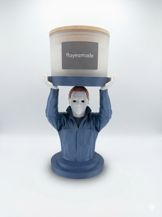 Masked Figure Candle Holder — navy blue bust of a figure in a zip-front coverall with a pale mask and both arms raised, holding a lidded frosted-white jar candle on a flat round platform, on a soft light-gray studio background.