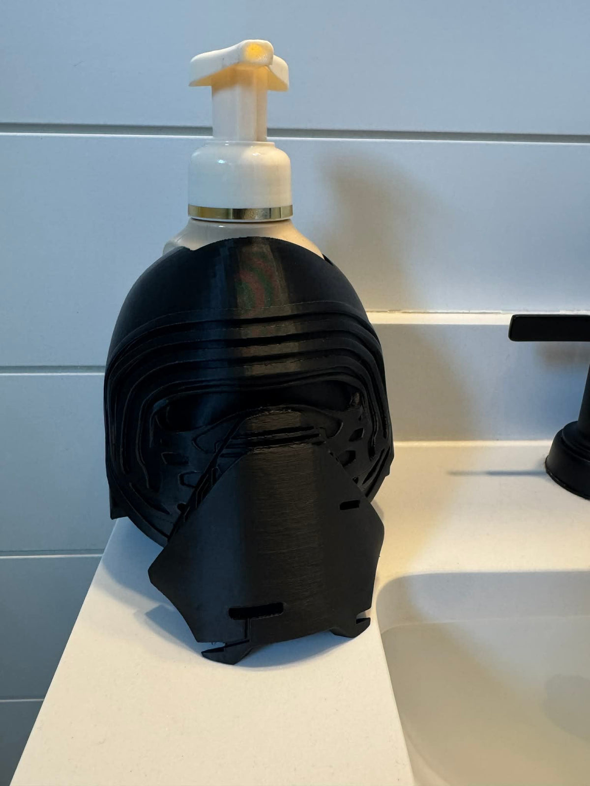 Masked Knight Soap Dispenser Holder — a matte black helmet-shaped holder on a bathroom counter, cradling a pump soap bottle.