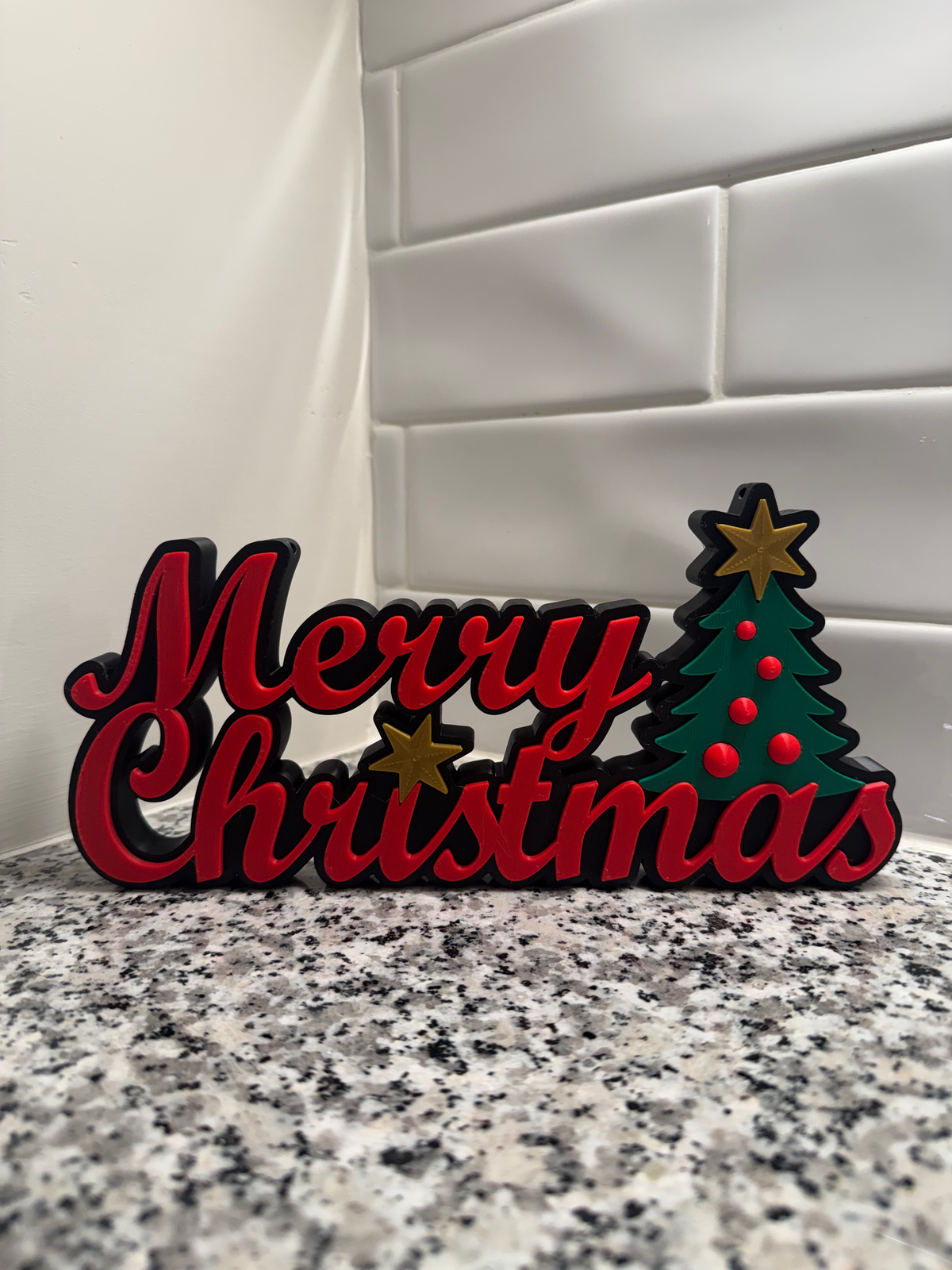 Merry Christmas Tree Sign — a red script 'Merry Christmas' sign with gold stars and a green decorated tree, standing on a granite counter.