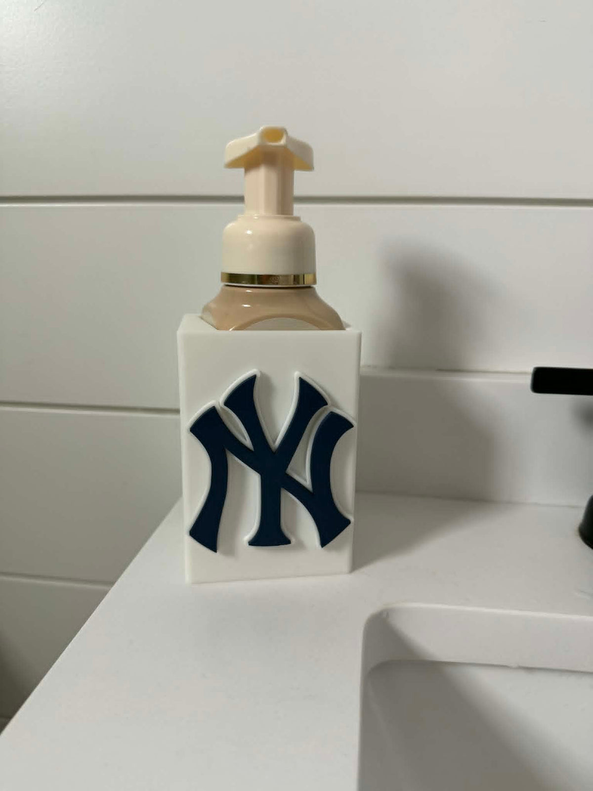 New York Soap Dispenser Holder — a white rectangular sleeve with a navy blue raised monogram, holding a foaming soap bottle at a bathroom sink.