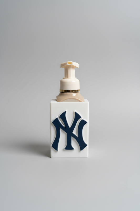 New York Soap Dispenser Holder — a white rectangular sleeve with a raised navy monogram, holding a foaming soap bottle with a cream pump, on a soft light-gray studio background.
