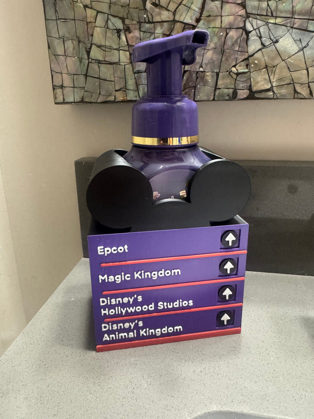 Park Street Sign Soap Dispenser Holder — a purple layered directional sign holder with four arrow panels and a rounded eared top piece, holding a purple foaming soap bottle on a counter.