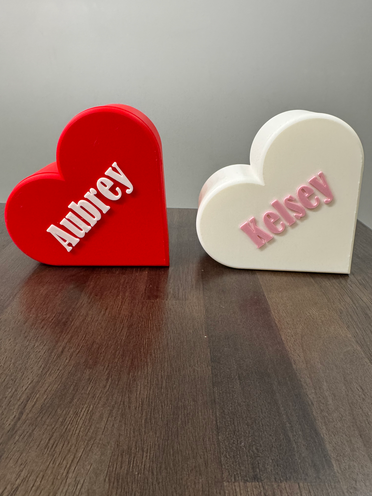 Personalized Heart Keepsake Box — two heart-shaped boxes, one red and one white, each with a raised personalized name on the lid, on a dark wood table.