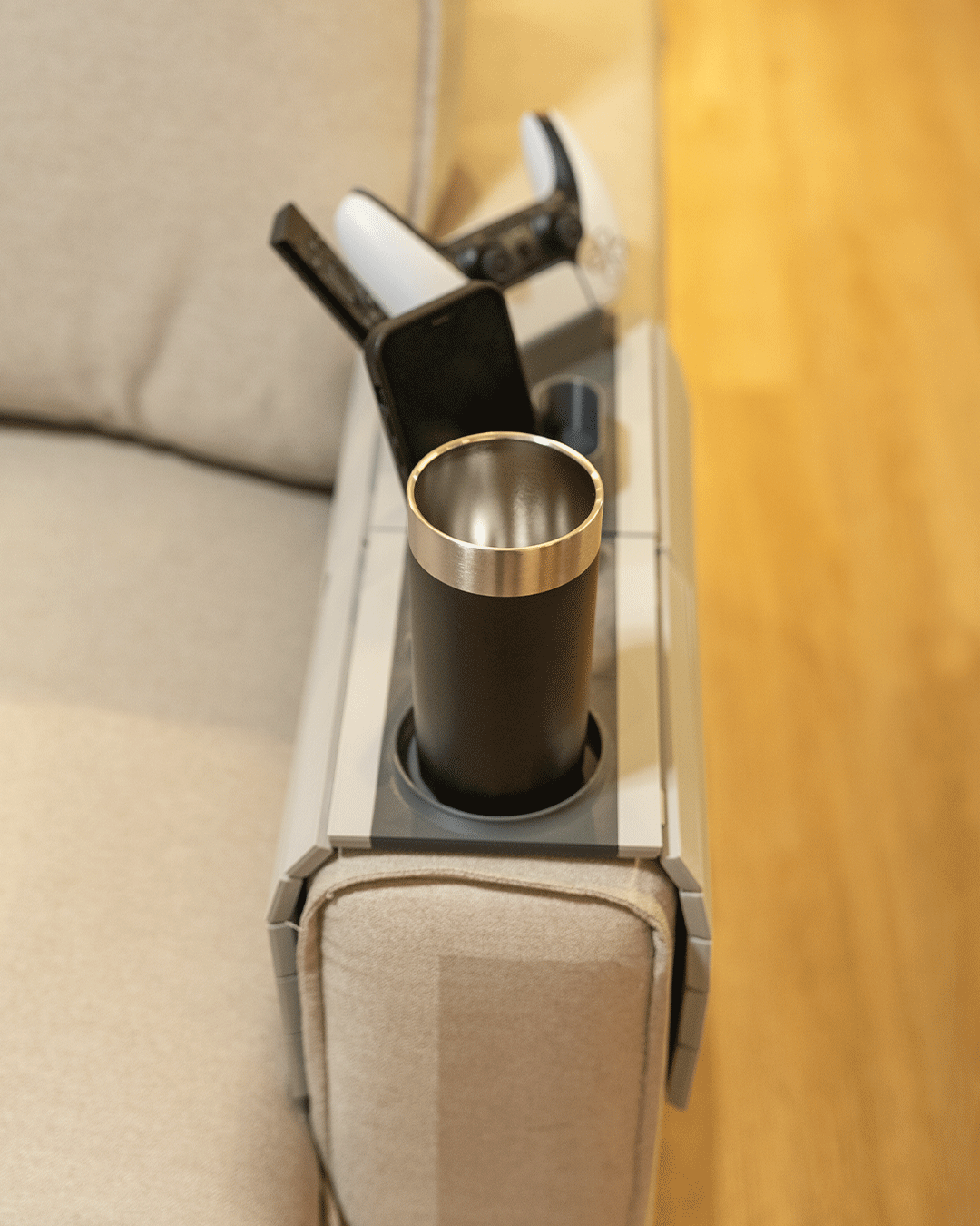 Player One Modular Sofa Side Table — angled view of the tray on a cream sofa arm with a tumbler in the cup holder and a controller behind it.
