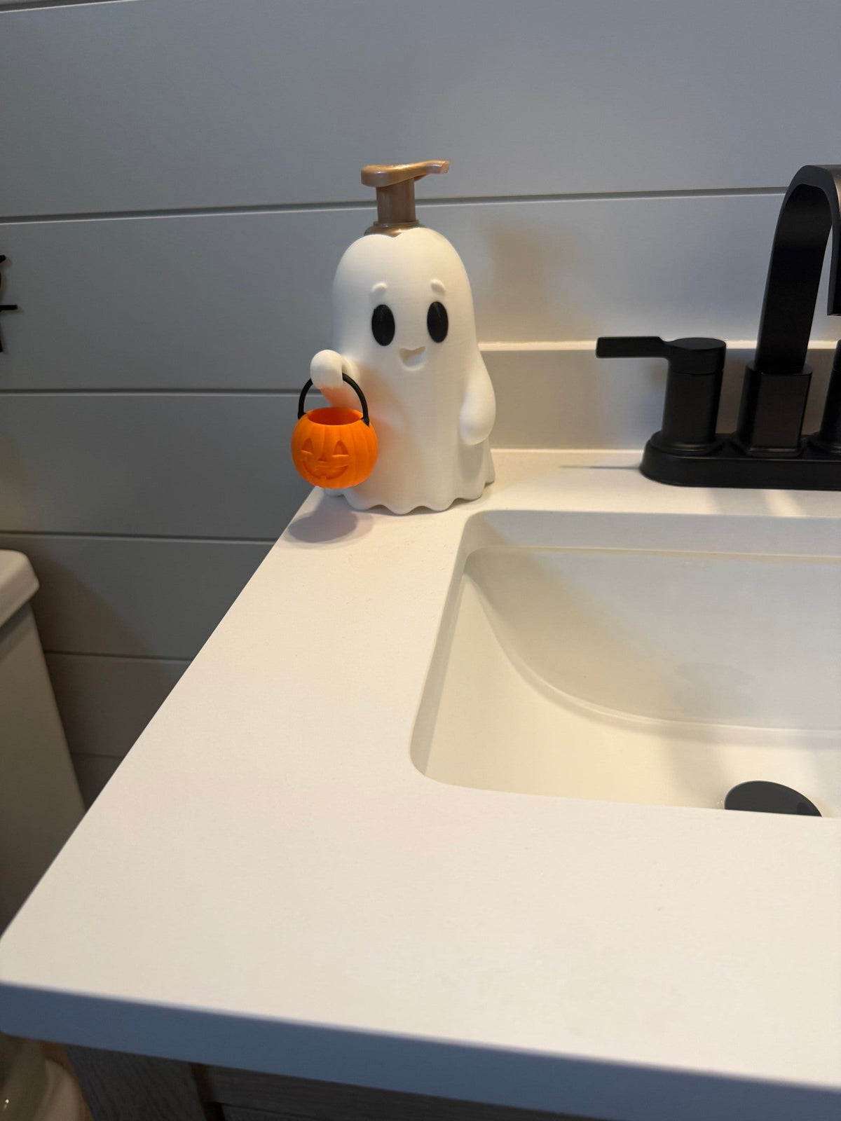 Pumpkin Pail Ghost Soap Dispenser Holder — a white ghost-shaped holder with a simple face carrying an orange pumpkin pail, holding a soap bottle at a bathroom sink.