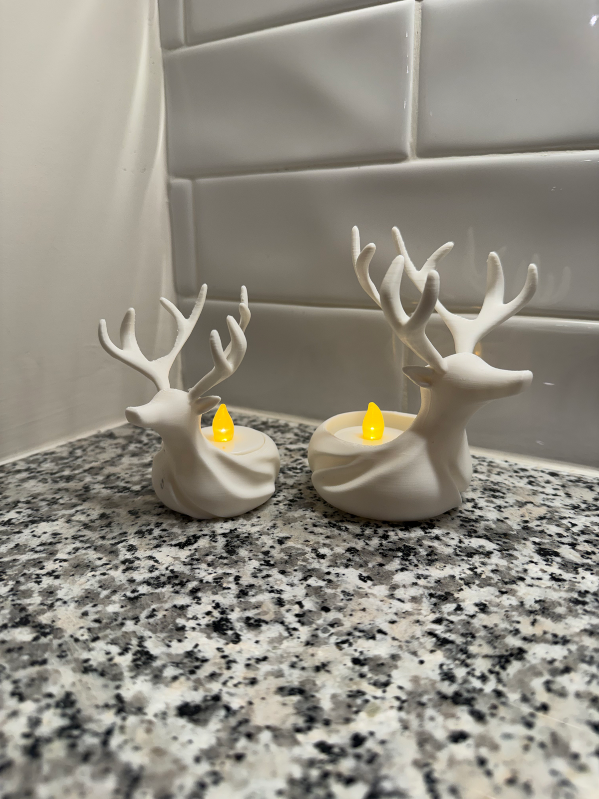 Reindeer LED Tealight Holders, Set of 2 — two white reindeer-shaped holders with antlers, each cradling a lit flameless tealight, on a granite counter.