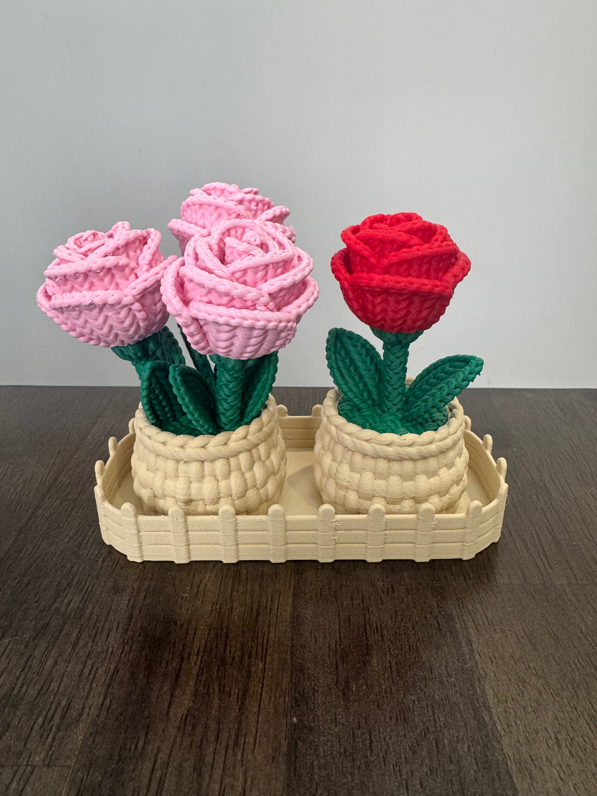 Rose Planter Tray Set — two woven-look basket planters holding pink and red roses with green leaves, sitting in a cream picket-fence tray on a dark wood table.