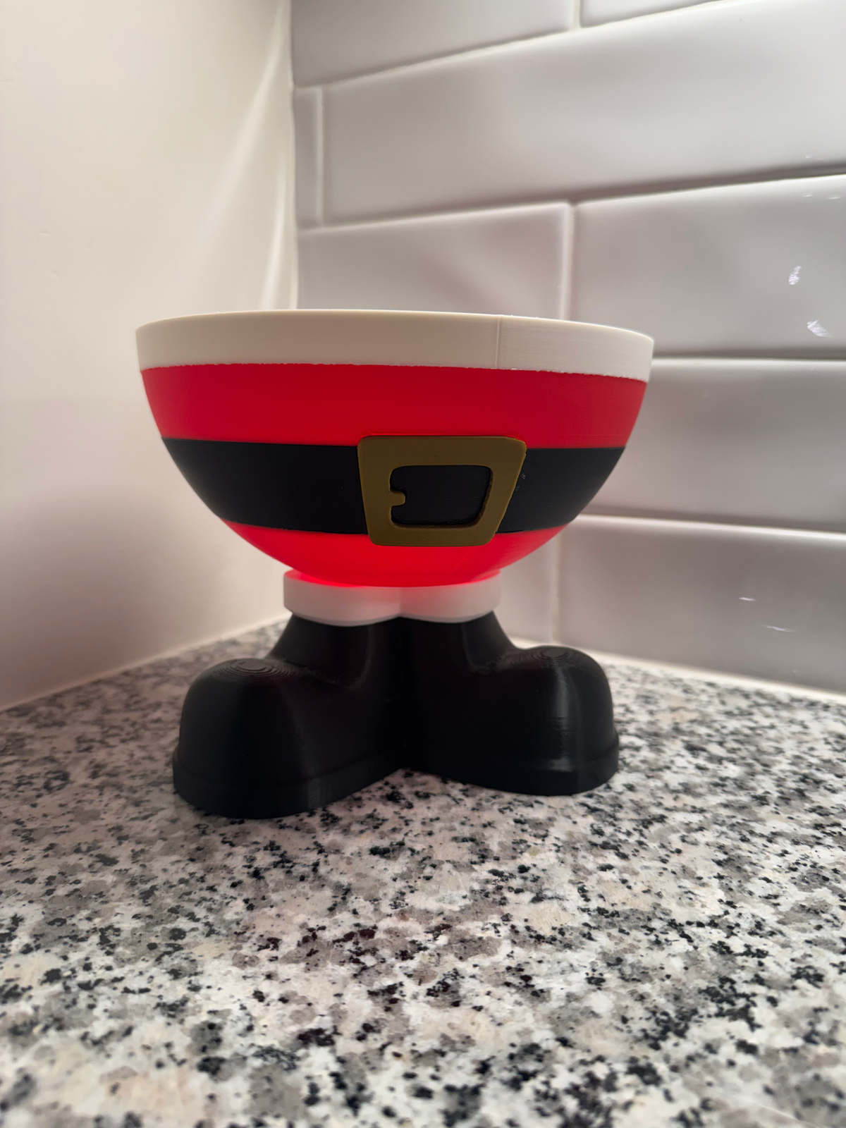 Santa Elf Trinket Bowl — a red and white bowl with a black belt and gold buckle, resting on black boot feet, on a granite counter.