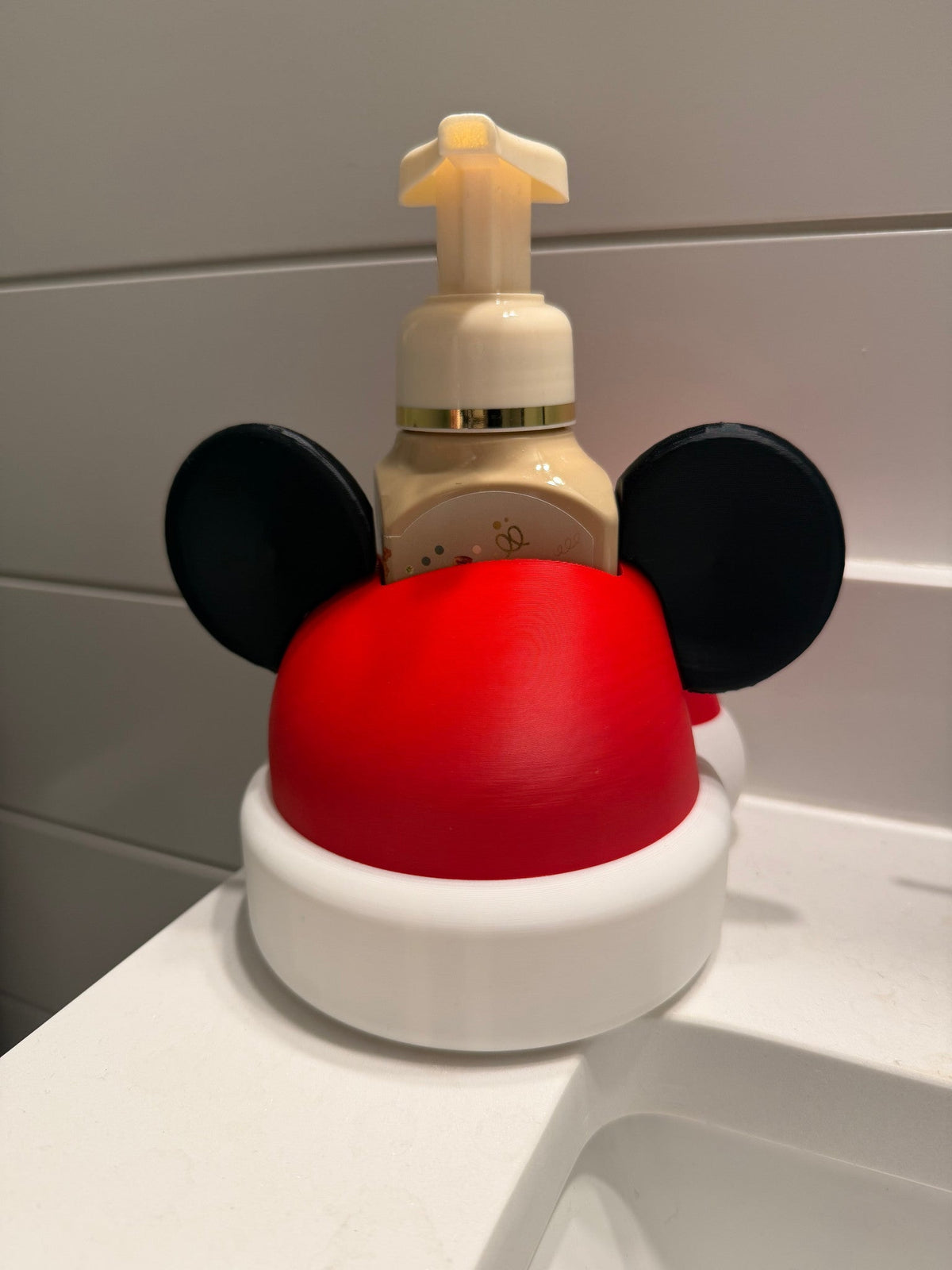Santa Hat Mouse Soap Dispenser Holder — a white base with black ear shapes and a red domed hat, cradling a foaming soap bottle at a bathroom sink.