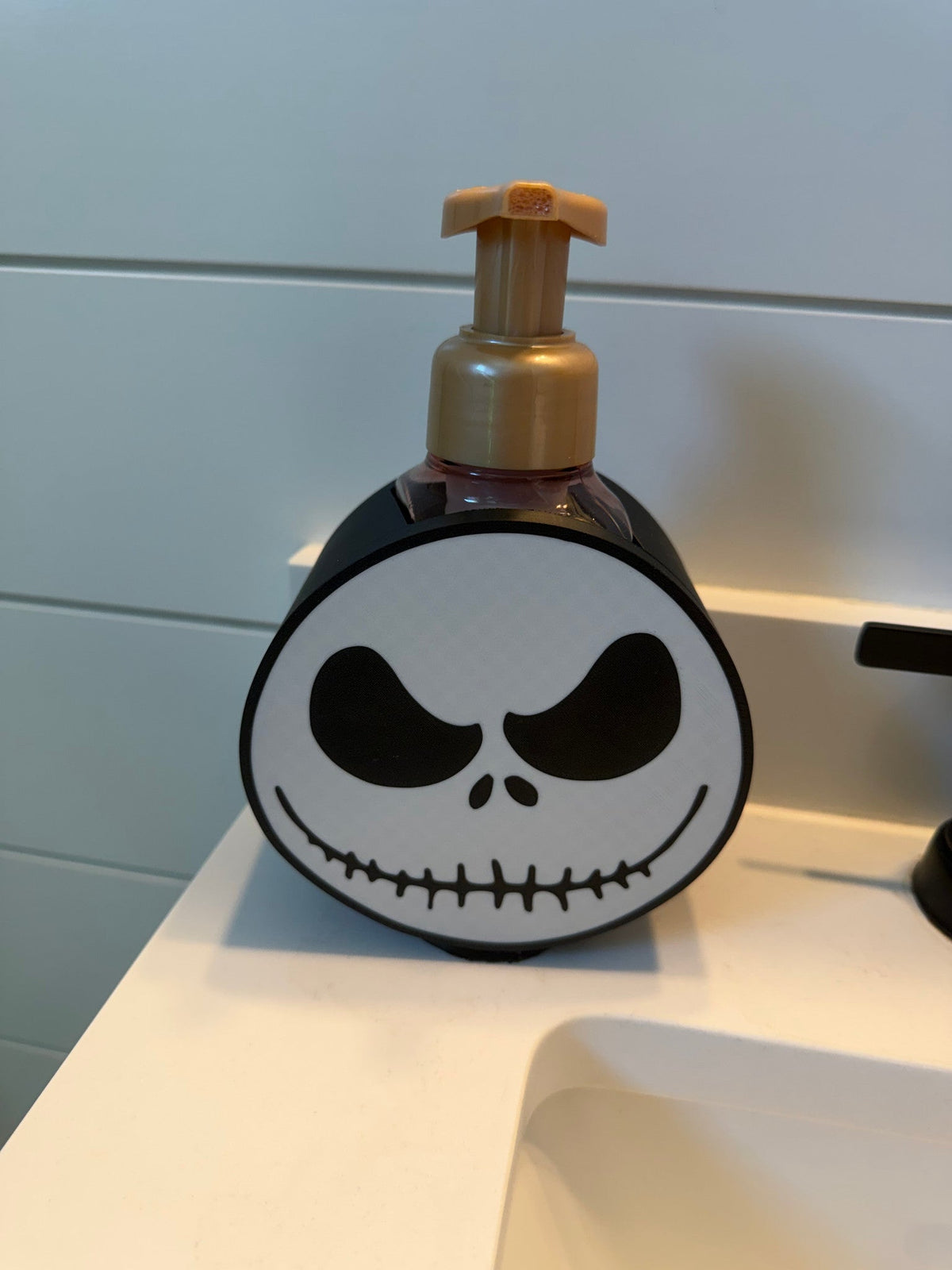 Skeleton Face Soap Dispenser Holder — a round black and white holder with a printed face design, cradling a dark soap bottle at a sink.