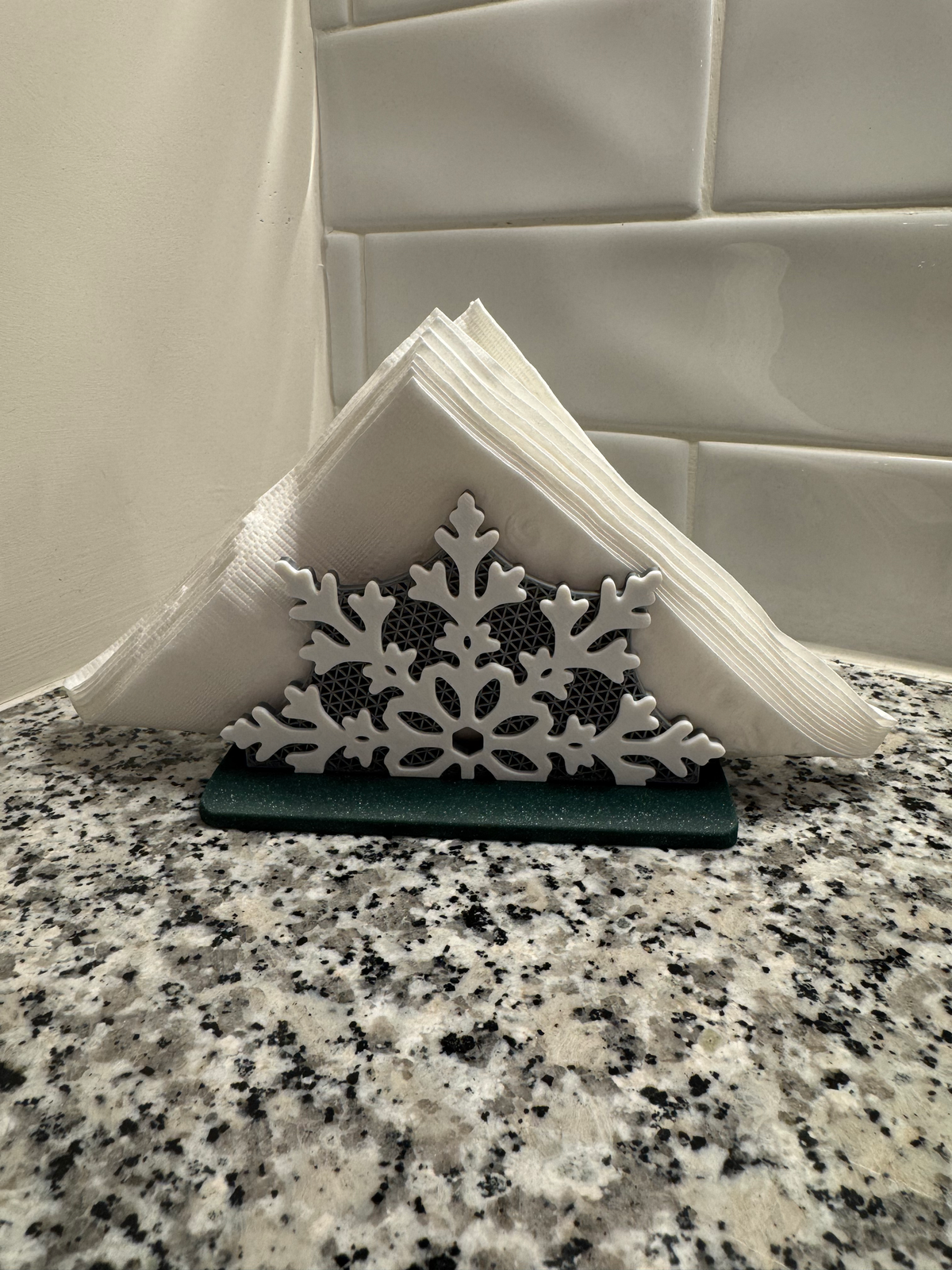 Snowflake Napkin Holder — a white snowflake-front napkin holder on a dark green base, holding folded napkins on a granite counter.