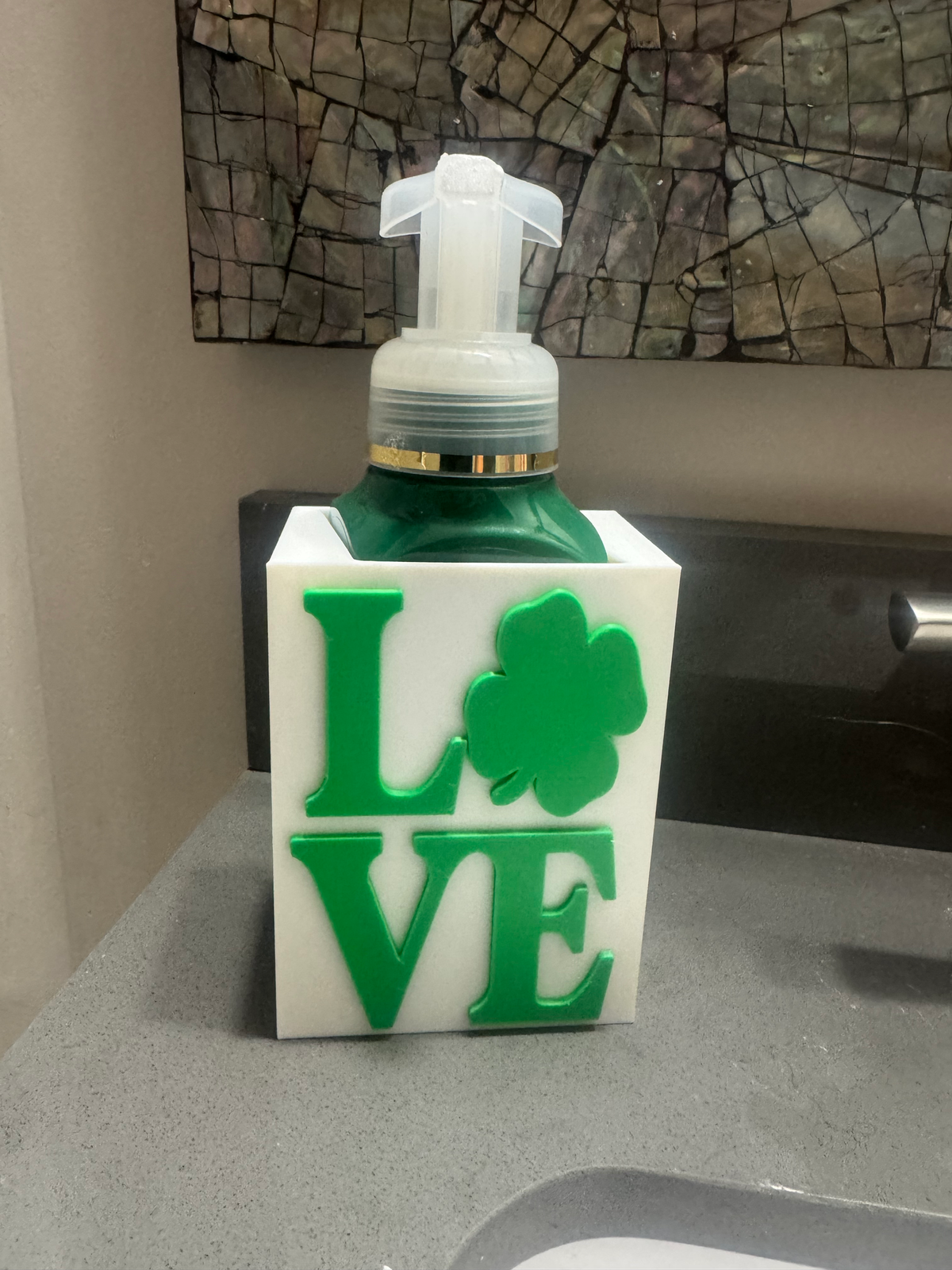 St. Patrick's Day LOVE Soap Dispenser Holder — a white square sleeve with raised green 'LOVE' lettering using a shamrock as the O, holding a green foaming soap bottle.