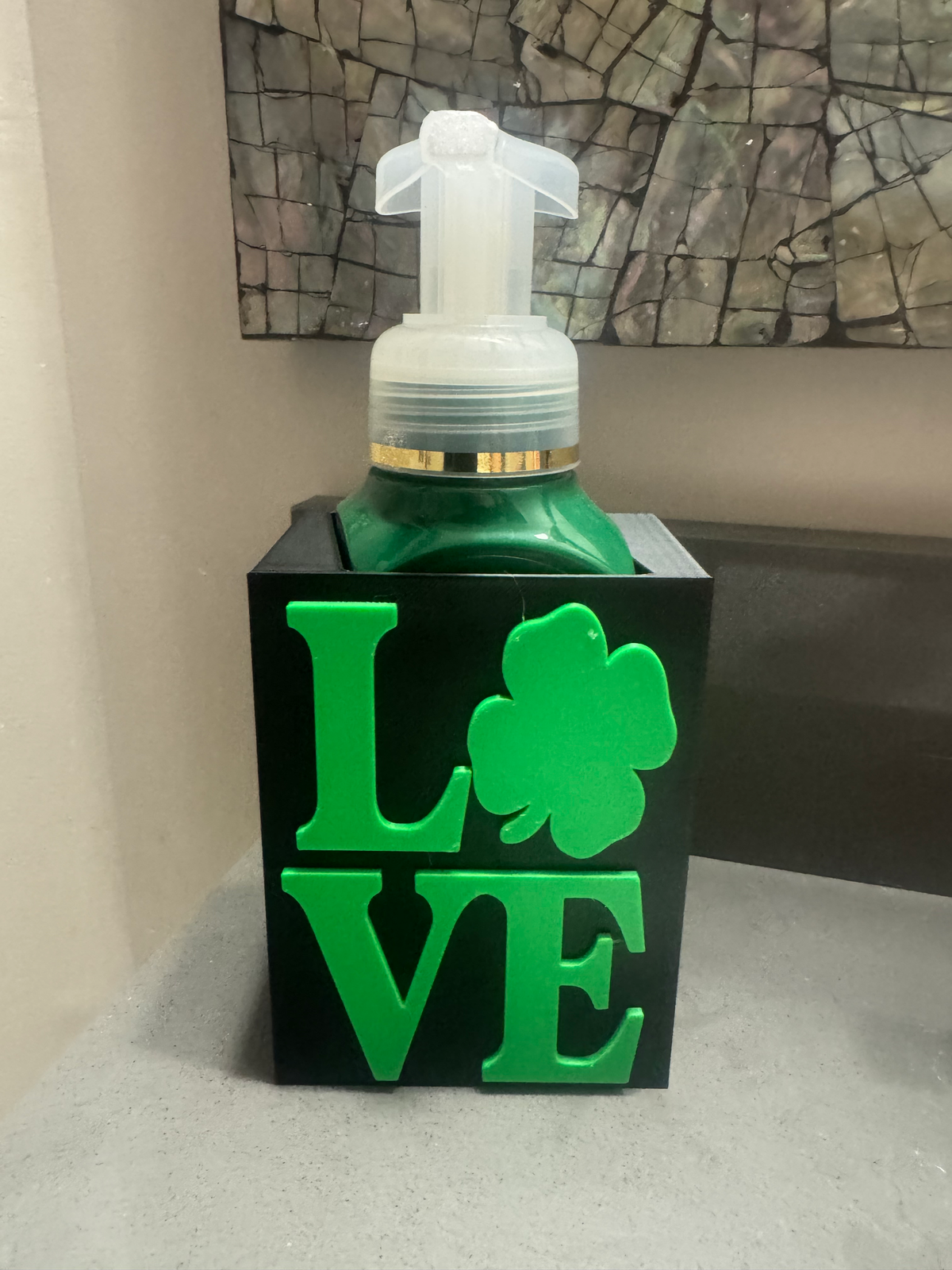 St. Patrick's Day LOVE Soap Dispenser Holder in black — a black square sleeve with raised green 'LOVE' lettering and a shamrock, holding a green soap bottle.