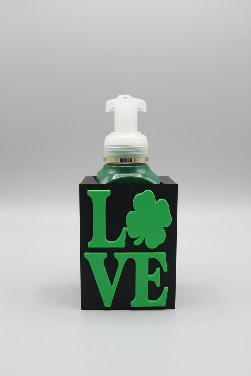 St. Patrick's Day LOVE Soap Dispenser Holder (Black) — a black rectangular base soap dispenser featuring the word LOVE in green and a green shamrock, filled with dark green liquid, on a soft light-gray studio background.