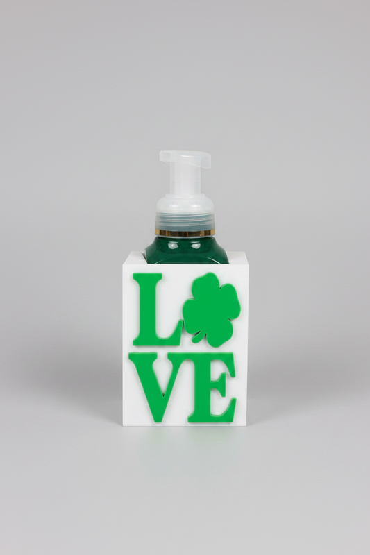 St. Patrick's Day LOVE Soap Dispenser Holder (White) — a white rectangular base soap dispenser featuring the word LOVE in green and a green shamrock, filled with dark green liquid, on a soft light-gray studio background.