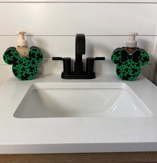 St. Patrick's Day Soap Dispenser Holder — a pair of black and green shamrock-patterned holders with ear shapes, one with a bow, flanking a black faucet on a white vanity.