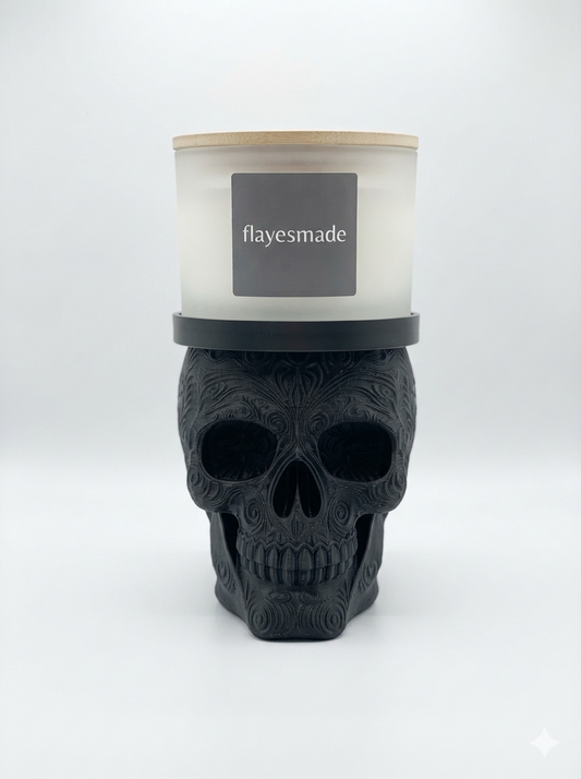 Sugar Skull Candle Holder — matte black skull with swirling filigree relief across the crown, brow, cheekbones and jaw, topped with a matching black collar ring holding a lidded frosted-white jar candle, on a soft light-gray studio background.