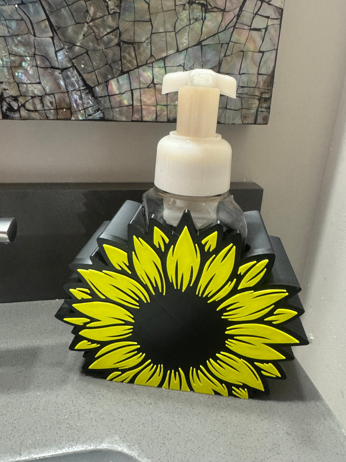 Sunflower Soap Dispenser Holder — a yellow sunflower with a black center mounted on a black stand, holding a foaming soap bottle on a bathroom counter.