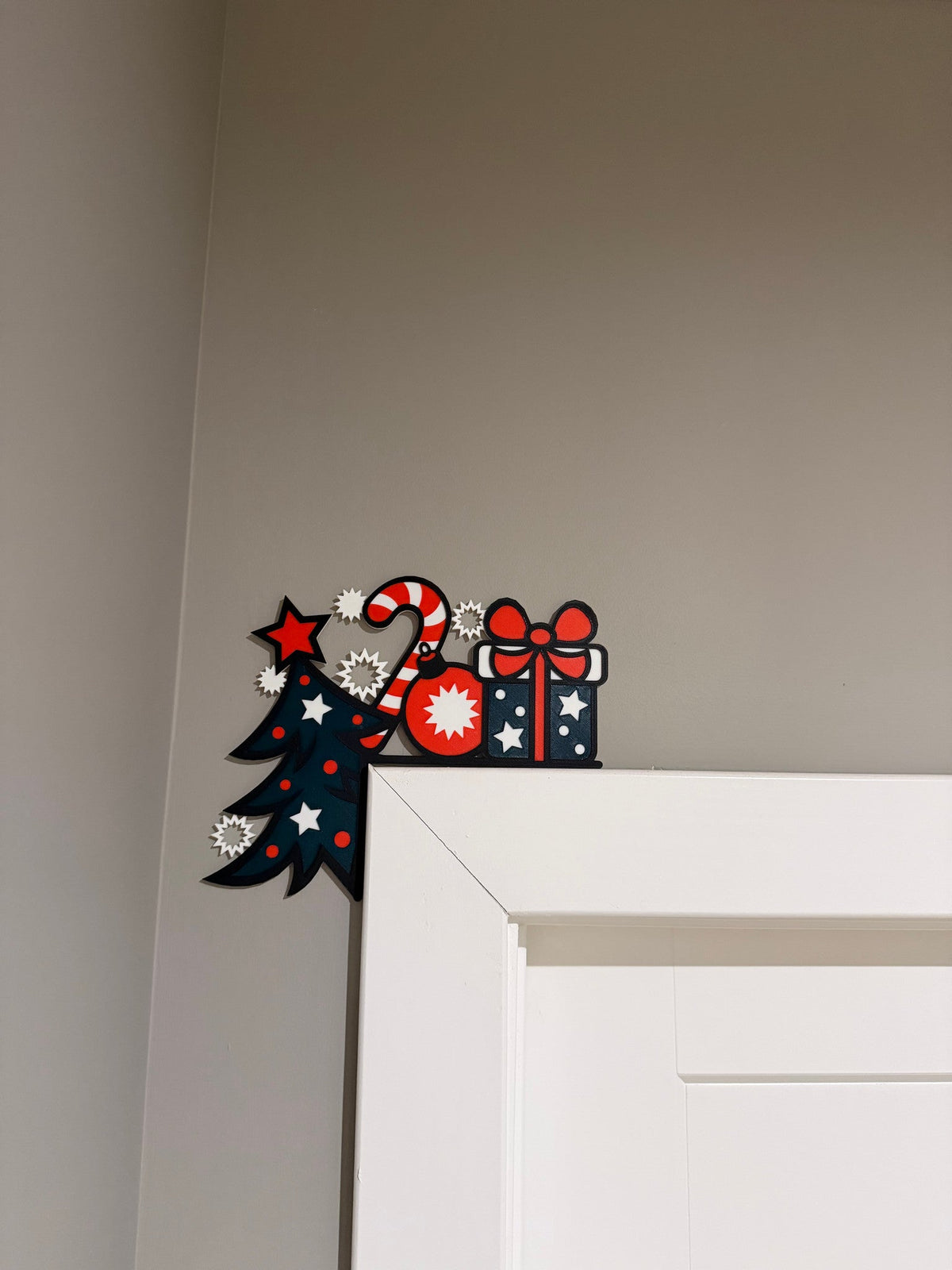 Tree Gift Candy Cane Corner Door Topper — a navy tree, candy cane and wrapped gift in red, white and blue, perched on the top corner of a white door frame.