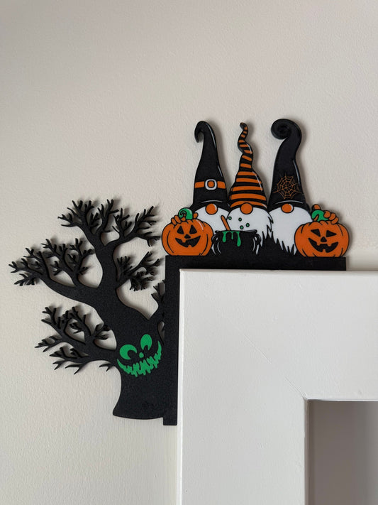 Witchy Gnome Trio Corner Door Topper — three gnomes in pointed black and orange hats with pumpkins, beside a bare black tree with a glowing green grin, on a white door frame.