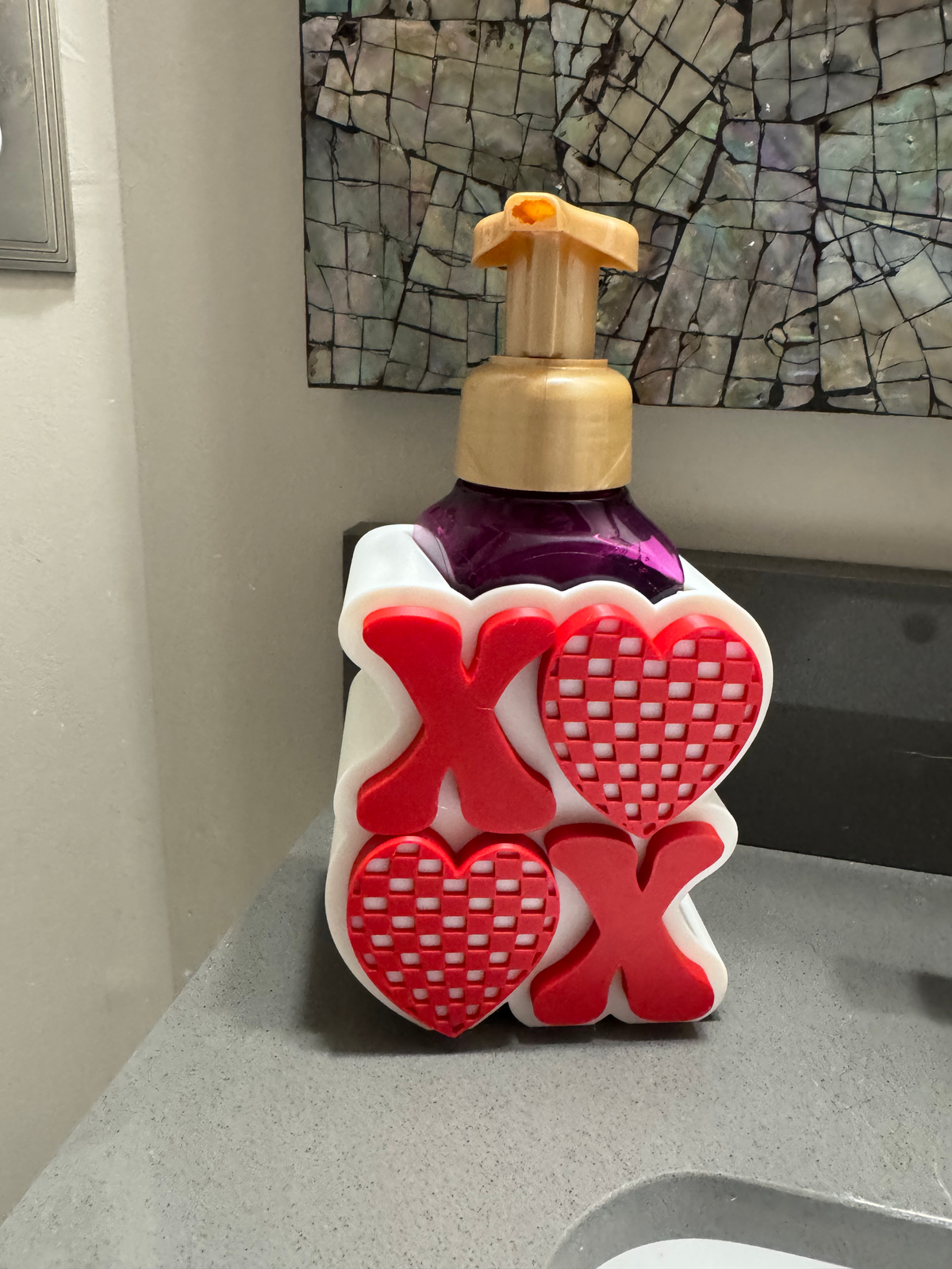 XOXO Heart Soap Dispenser Holder — a white sleeve with raised red X's and checkered hearts, holding a purple foaming soap bottle on a counter.