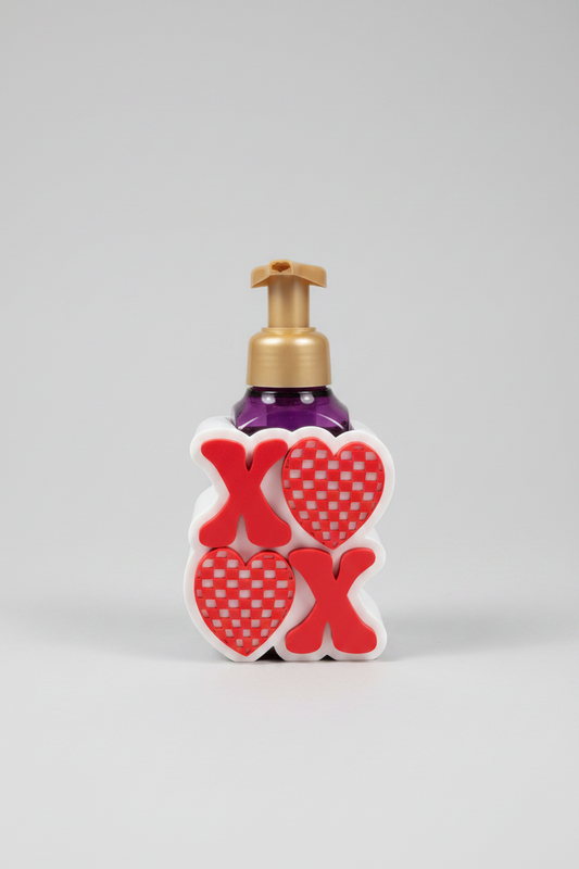 XOXO Heart Soap Dispenser Holder — a Valentine's Day themed soap dispenser shaped like a white block with red and red-and-white checkered hearts, featuring a gold pump and a purple bottle, on a soft light-gray studio background.