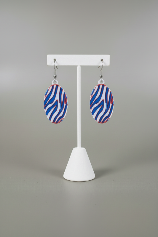 Zigzag Stripe Earrings — Buffalo Colors — a pair of oval drop earrings with a red, white, and blue zigzag pattern, hanging from silver fishhook wires, on a soft light-gray studio background.